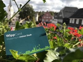 Die WippCard, im Hintergrund sind rote Blumen mit grünen Blättern und mehrere Gebäude am Marktplatz