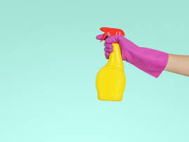Bild mit hellblauem Hintergrund. Eine Hand in einem lila Handschuh hält eine gelbe Sprühflasche